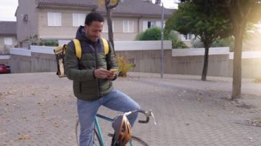 Yemek teslimatı konsepti. Sırt çantalı genç siyahi adam şehirde akıllı bir telefon ile online alışveriş siparişi veren müşterilere ürün dağıtıyor. Siyah erkek işinden memnun..