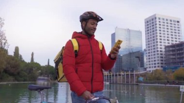Yemek teslimatı konsepti, bisikletle yiyecek getiren mutlu Afrikalı Amerikalı genç binici. Gülümseme şehirdeki akıllı telefonunun konumunu kontrol ediyor. Yüksek kalite 4k görüntü