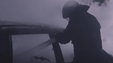 Unrecognizable fireman silhouette combating a fire of a burning car accident at night. Firefighter fighting flames with water hose. Filmed with RED camera. High quality 4k footage