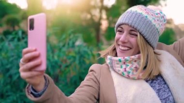 Young beautiful woman smiling taking selfie with pink smart phone in winter. Trendy blonde girl in casual attirePositive cheerful female student posing outdoors for sharing in social media app
