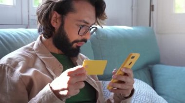 Young man shopping online using credit card and phone on the sofa at home. Male ordering online successful payment using smartphone. High quality 4k footage