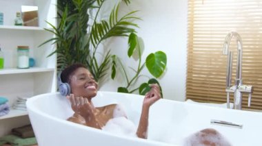 Attractive black woman wearing headphones happy relaxing and dancing in foam bath in beautiful bathroom with plants. Beauty, skin care and wellbeing concept. 4k footage shot on RED camera slow motion.