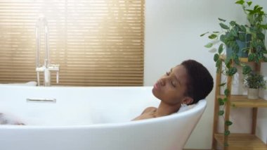 Attractive woman relaxing in foam bath in beautiful bathroom with plants at home during a sunny morning routine. Calm, pleasure, happiness and wellbeing concept. 4k shot on RED camera slow motion