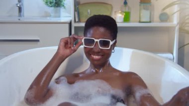 Portrait of an attractive African woman in foam bathtub with sunglasses looking at camera smiling happy. Cool funky lifestyle. Real people wellness concept. 4k shot on RED camera in slow motion