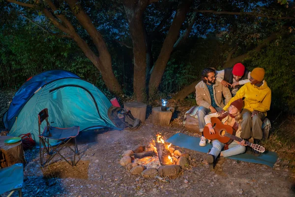 Group of friends with guitar near the bonfire and camping outdoors in the afternoon. Charismatic colleagues sitting by the fire laughing and singing. Travel concept.