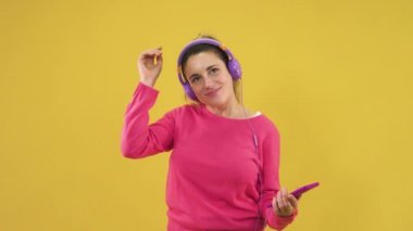 Kulaklıklı ve akıllı telefon dansı yapan bir kadın sarı arka planda izole edilmiş mutlu bir şekilde. Kulaklıklı beyaz kadın müzik dinliyor..