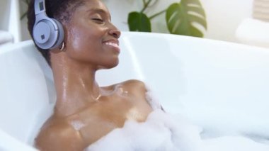 Kulaklık takan çekici Afrikalı kadın köpüklü küvette müzik eşliğinde dinleniyor. Güzellik, cilt bakımı, spa ve refah konsepti. Yüksek kalite 4k görüntü