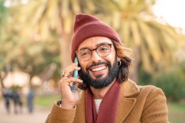 Attractive young caucasian man with glasses and beard talking in the phone outdoors in the park in winter. Happy smiling student call. Technology, education and communication concept.