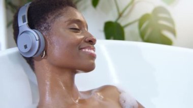 Kulaklık takan çekici zenci kadın köpüklü küvette müzik eşliğinde rahatlıyor. Güzellik, cilt bakımı, spa ve refah konsepti. Yüksek kalite 4k görüntü