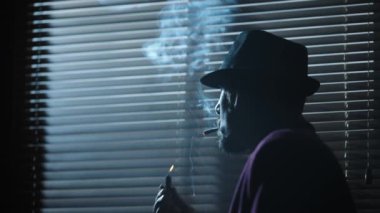 Kara film. Karanlık bir odada sigara içen şapkalı bir adam pencereden bakıyor. Yüksek kalite 4k görüntü