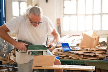 Zanaatkar, çeşitli malzemelerle dolu parlak bir atölyede zımpara makinesiyle pürüzsüz ahşabı rafine eder.