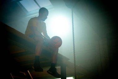 Elinde topla oturan genç bir basketbolcunun silüeti dramatik ışıklandırma efektleriyle çevrili..