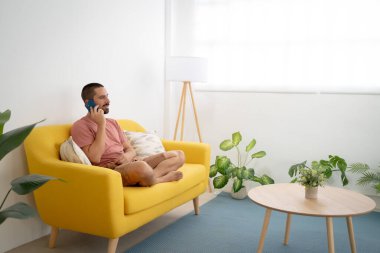 Gülümseyen genç adam evdeki koltukta oturmuş akıllı telefondan konuşuyor. Yüksek kalite fotoğraf