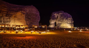 Illuminated outdoor lounge in front of elephant rock erosion monolith standing in the night desert, Al Ula, Saudi Arabia clipart