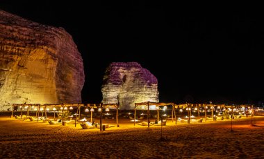 Illuminated outdoor lounge in front of elephant rock erosion monolith standing in the night desert, Al Ula, Saudi Arabia clipart