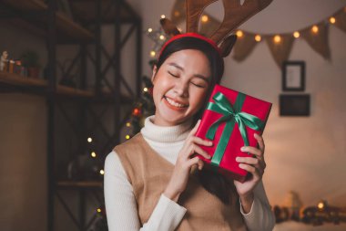 Süslü saç bandı takmış genç Asyalı bir kadın gülümsüyor ve elinde kırmızı bir kutuda bir kurdele ve arka planda süslü bir Noel ağacı olan oturma odasında bir Noel hediyesi tutuyor..