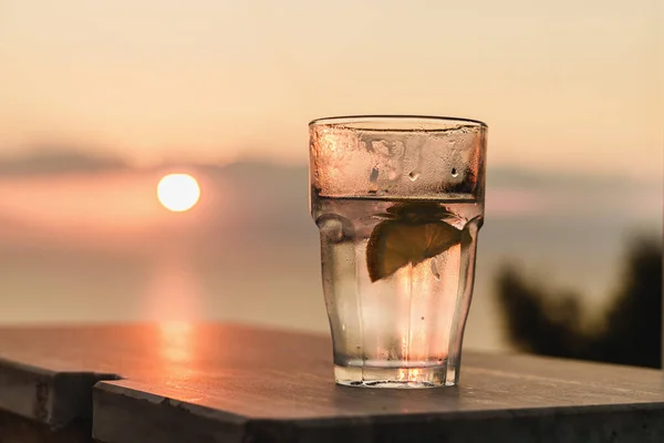 Fresh iced glass with transparent long drink - such as vodka tonic or gin tonic - with a slice of lemon outdoors at sunset against sun and cloudy sky background - drink beverage alcohol concept