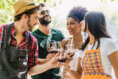 Joyful multiracial friends at picnic celebrating toasting with red wine standing under the trees in the nature - wine cheers - multi-ethnic people, food and drink lifestyle concept