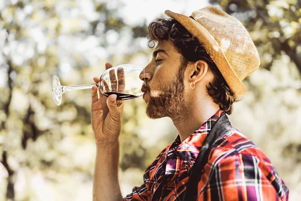 Handsome bearded guy drinking red wine at the grape harvest picnic under the trees and vines - food and drink - people lifestyle concept