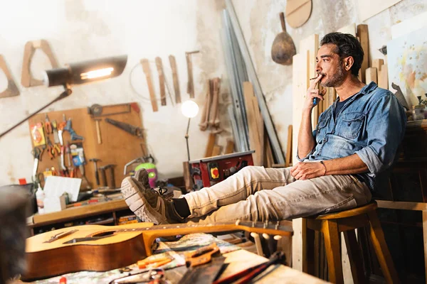 A carpenter relaxes sitting on a stool by placing his feet on the work table of his vintage woodwork - small business jobs, work and smoking addiction lifestyle concept