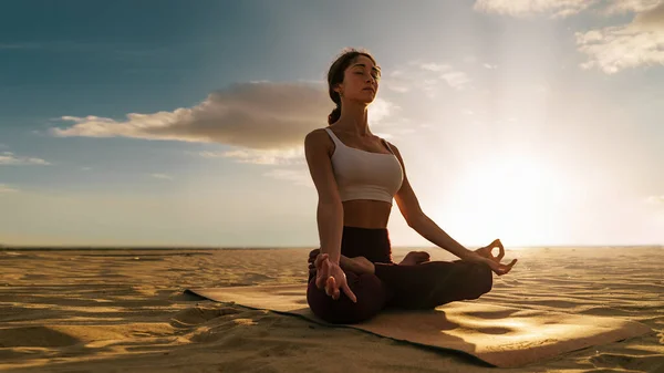 Young woman meditating in lotus yoga pose on beach - peaceful scene of a girl making mindfulness exercises eyes closed sitting on the sand - people and spirituality lifestyle concept