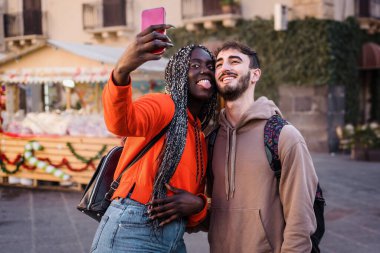 Engaged man and woman of different ethnicities enjoying a road trip in the city center. Interracial couple pose for a selfie holding each other with evening sun in the background.