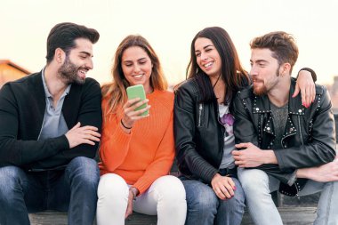 Happy group of trendy young people laughing and chatting sitting in a bench at sunset using a cellphone. Millennial friends having fun together outdoor - people, social network lifestyle concept
