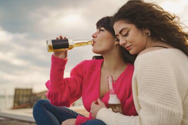 Two young caucasian women sit on a city wall, drinking Mexican beers and embracing each other, one with smooth hair, the other with curly hair, while a cloudy and gray sky serves as a background