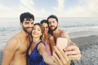 Dört genç insandan oluşan bir grup, iki erkek ve iki kadın, arkalarında okyanus ufkuyla sahilde selfie çekiyorlar. Hepsi mayo giyiyor ve birlikte harika vakit geçiriyor gibi görünüyorlar.