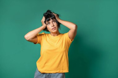 A portrait of a young, caucasian brunette woman expressing stress by pulling her hair and looking upwards. She is wearing jeans and a red t-shirt and is against a green background.