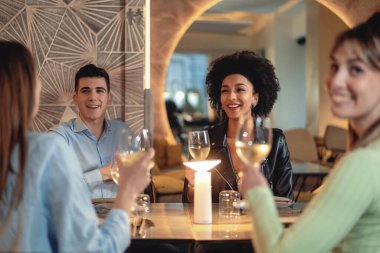 A group of four friends sits at a restaurant and happily chat while holding a glass of white wine. The focus is on a young African American woman with curly hair who is smiling