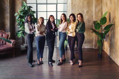 Six diverse women of various ethnicities and ages, including Asian, Eastern European, Afro-American, and European, smiling and posing with crossed arms in a business portrait photo taken in an office setting clipart