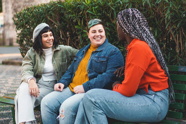 Three women, two Caucasian (one brunette, one curvy and non-binary) and one African with dreadlock-like extensions in her hair, sit together on a park bench, engaged in conversation