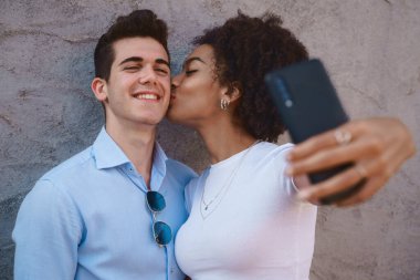 Multiracial couple taking a selfie, a young African American woman with curly hair kissing a Caucasian man on the cheek against a cement wall. Love, interracial, diversity, relationship concepts