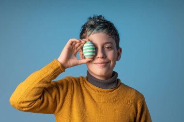 Happy 10-year-old caucasian boy with blue hair covering one eye with a decorated Easter egg on a blue background. Perfect for Easter-themed designs or illustrating children's crafts and activities