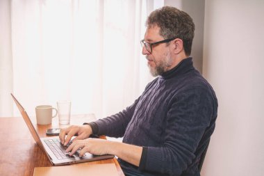 A middle-aged Caucasian man with grey beard is sitting at a desk and typing on a laptop computer. He is wearing a blue turtleneck sweater and glasses and is focused on reading the screen and typing on the keyboard