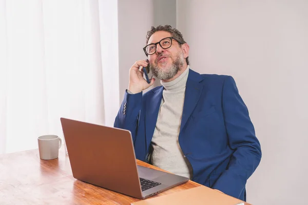 Mature businessman with beard and glasses talking on phone while sitting at desk with laptop computer. He is wearing a white turtleneck sweater and blue jacket