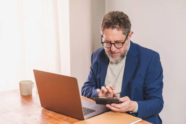 身着休闲装 头戴高领毛衣 身穿蓝色夹克 留着胡子和眼镜的中年商人 坐在办公室桌上 用一台大型计算器进行计算 — 图库照片