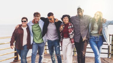Bir grup çok kültürlü arkadaş deniz kenarında yürüyorlar, kollarını birbirlerinin omuzlarına dolayıp gülüyorlar ve iyi vakit geçiriyorlar. Arka planda deniz görünüyor..