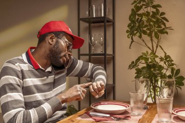 Gündelik kıyafetleri ve kırmızı şapkası olan genç Afrikalı bir adam, boş bir sandalyeyle iki kişilik bir restoran masasında oturuyor, telefonla konuşuyor ve kol saatini işaret ediyor, dakikliği teşvik ediyor.