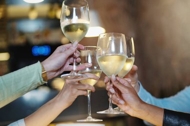 Koyu tenli biri de dahil olmak üzere çeşitli ellerin yakın plan ayrıntıları bir barda ya da gece aydınlatmalı bir restoranda beyaz şarapla kadeh kaldırıyor..