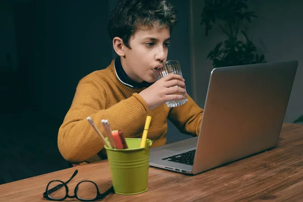 彼の机に座っている若い男の子は ガラスから水を吸いながらノートパソコンに集中し 鎮静作用を発揮しながら水分補給を続けることの重要性を強調しています — ストック写真