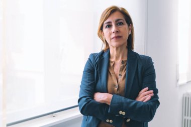 50 'li yaşlarda, beyaz ofis odasının penceresinin yanında duran, sıradan ofis kıyafetleri içinde kolları çaprazlanmış, ciddi bir şekilde kameraya bakan beyaz bir iş kadını..