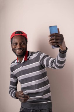 Moda kıyafetli ve şapkalı genç adam temiz bir stüdyo ortamında akıllı telefonuyla selfie çekiyor. Modern yaşam tarzı ve teknoloji temaları için mükemmel..