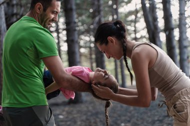 Baba kızını ağaçların arasında tutarken anne onu sevgiyle okşuyor. Ormanın kalbinde yürekten bir sahne.