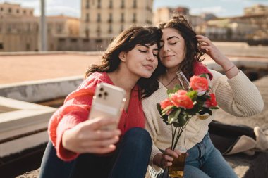 20 'li yaşlarda genç lezbiyen bir çift rıhtımda selfie çekerken gözleri kapalı birbirlerine yaklaşırlar. Biri bir buket kırmızı gül tutuyor..