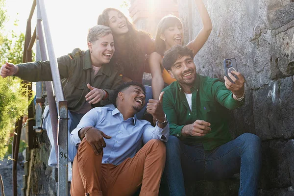 A group of five people, including a non-binary person and an African descendant, are at the seaside steps, capturing a joyful selfie together with a smartphone.