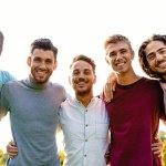 Um grupo multirracial de cinco amigos do sexo masculino abraça ao ar livre, compartilhando um momento de unidade e alegria. Todos sorriem calorosamente, dirigindo suas expressões alegres para a câmera.