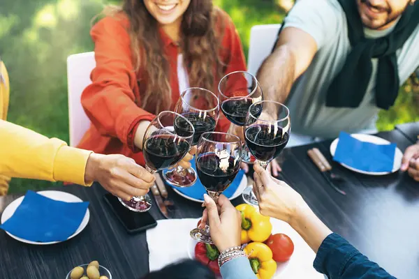 A close-up detail of wine glasses clinking in a toast on a courtyard table. The blurred and out-of-frame faces of the young friends hint at the joyous gathering.