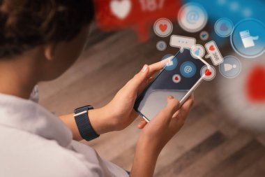 Arkadan görülen genç bir çocuk, akıllı telefonunu kullanıyor. Cihazdan sosyal ağ etkileşimlerini temsil eden grafiksel olarak tasarlanmış simgeler - dijital dünyanın bağlanabilirliği ve bağlılığı.